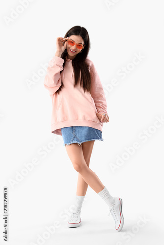 Stylish young woman in pink hoodie with sunglasses on light background