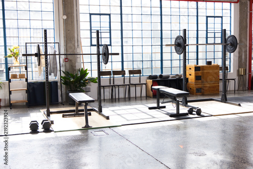 Two bench press stations are occupying raised platform in sunlit warehouse gym, with barbells