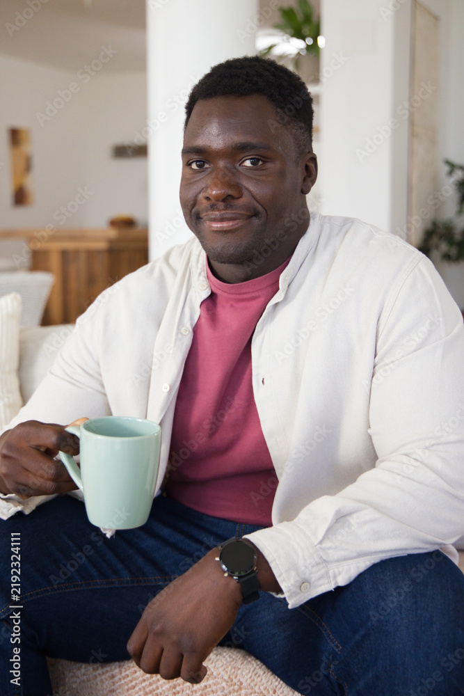 Fototapeta premium African American man wearing maroon shirt sitting on sofa in living room holding mint-green mug