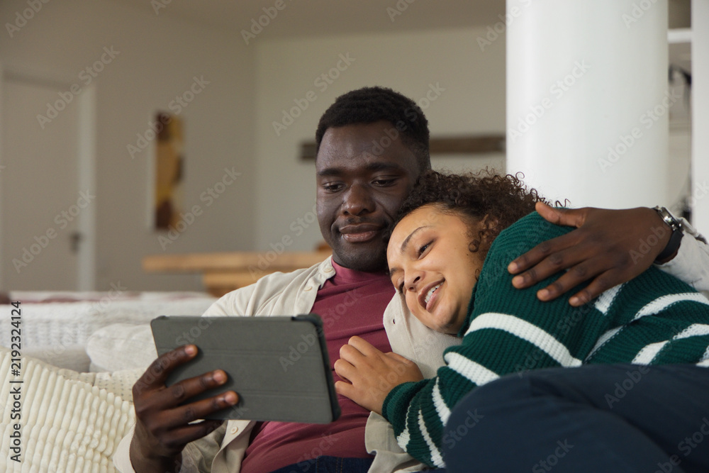 Obraz premium African American couple sitting close on light-colored sofa at home, watching tablet screen
