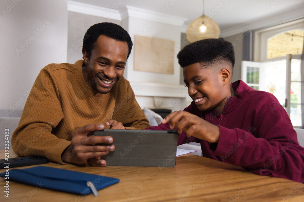Fototapeta premium African American father and teenage son leaning and tapping tablet in living room with papers