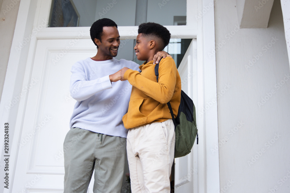 Obraz premium African American father with teen son sharing fist bump at doorway, backpack visible, white door