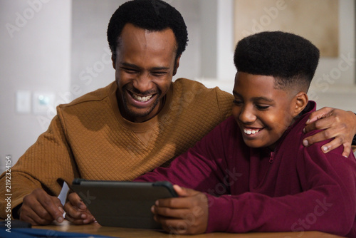 African father, teen son sharing tablet in black case at wooden table at home, maroon zip-up