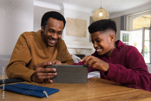 African American father and teenage son leaning and tapping tablet in living room with papers