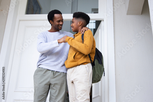 African American father with teen son sharing fist bump at doorway, backpack visible, white door