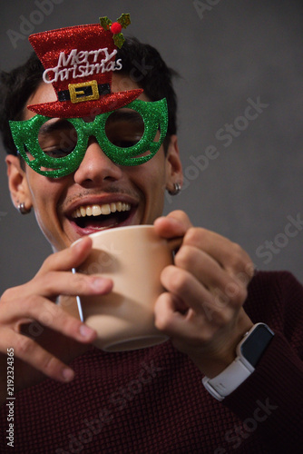 Adult African American man wearing glitter Merry Christmas glasses, holding beige mug, smiling