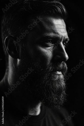 Close up portrait of serious bearded man in dark studio. Mature caucasian male with thick beard looks away with intense gaze. Dramatic low key lighting creates shadow on face. Mysterious moody
