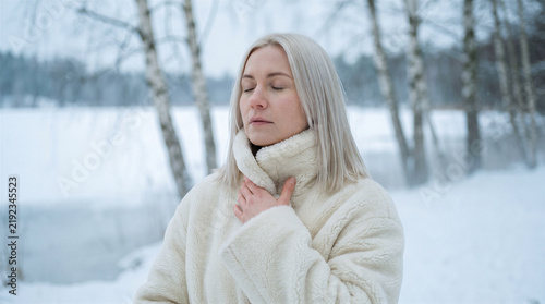 Woman with blonde hair wearing a white fleece coat standing in a snowy winter forest with eyes closed