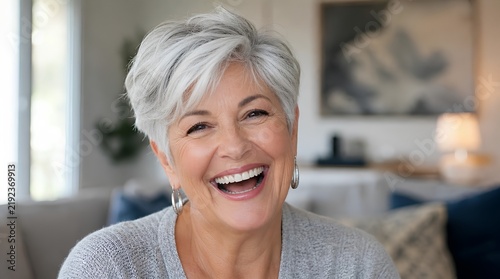 Close-up portrait of a happy elderly woman with a stylish short silver haircut laughing openly in a bright modern living room interior with soft natural light