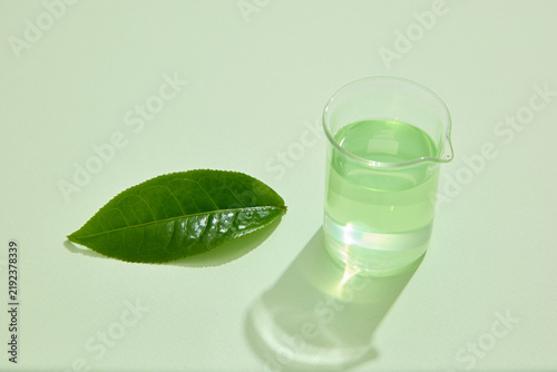 A single green tea leaf beside a clear glass container on a pale green background, creating a minimal skincare ingredient composition with copy space.