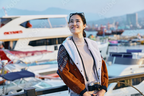 Fotografie Woman in a cozy jacket with sunglasses by a busy marina, smiling, holding a drin