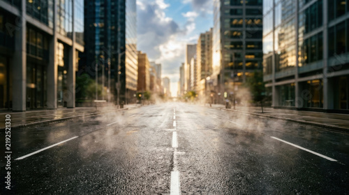 Empty city street with wet asphalt and steam rising between modern skyscrapers during early morning