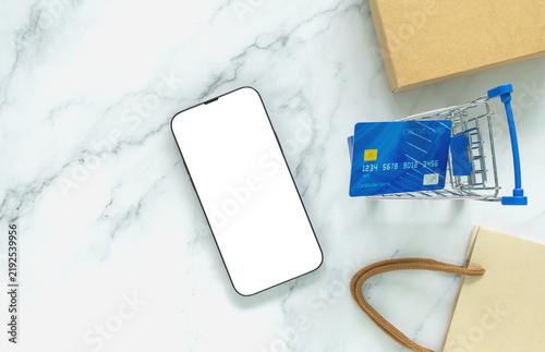 Blank smartphone mockup with credit card, shopping cart and kraft package on white marble, illustrating ecommerce, online shopping, mobile payment, retail, digital commerce and product promotion.