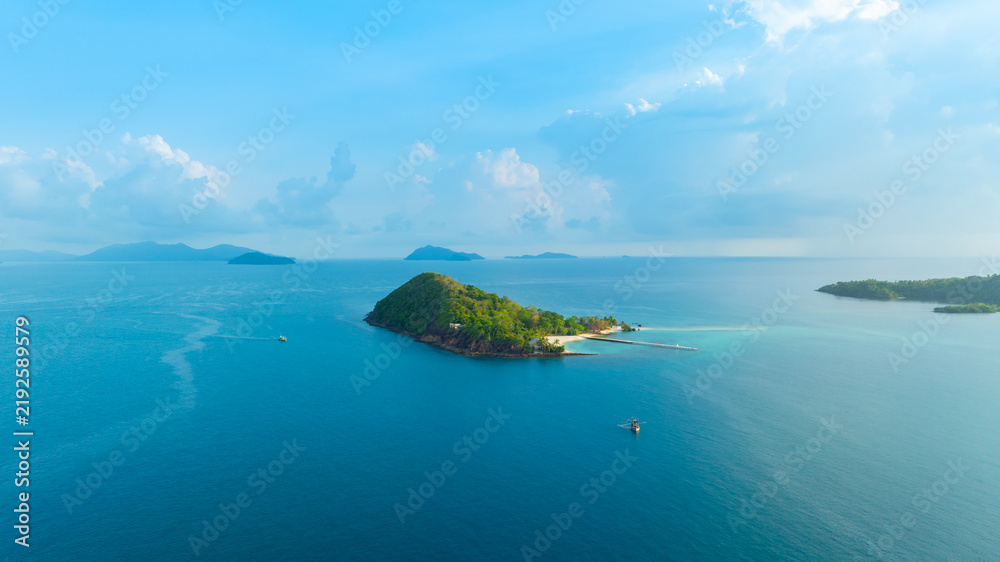 Obraz premium Panoramic aerial view of a lush green tropical island in the middle of a vast blue ocean under a bright sky with clouds, representing vacation, travel, and scenic nature.concept travel