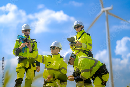 Wind farm crew of four in yellow and navy coveralls talk over a small drone while the man in front straightens up from a crouch and a turbine rotor blurs behind them