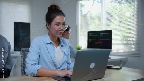 Asian female programmer developing software code on a laptop computer with her team in an office