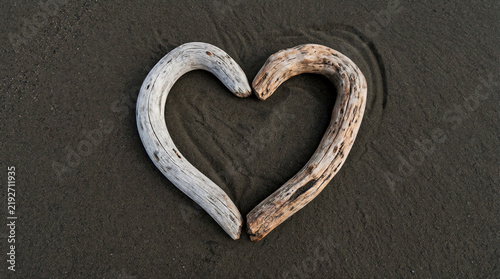 Heart shape formed by two smooth driftwood pieces, one worn smooth and one rough, resting on dark sand