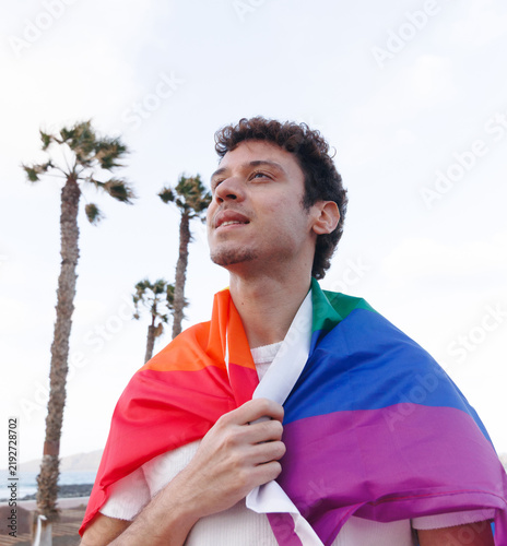 A young man proudly walks along the seashore carrying the LGBTQ flag