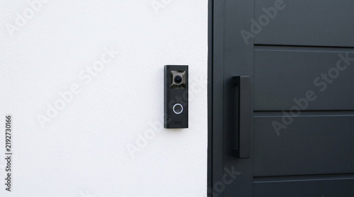 Sleek smart video doorbell mounted on a white textured wall next to a modern black door, representing high-tech home security and automation.