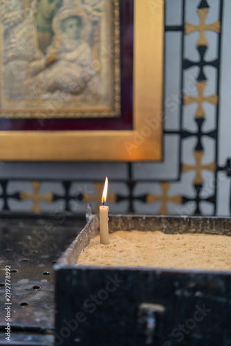 View of a single burning candle placed in a sand-filled metal container inside a church in Coptic Cairo, with a blurred religious icon in the background, Cairo, Cairo Governorate, Egypt.