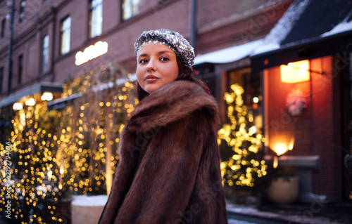 Young woman with dark hair wearing a fur coat and sequined headband stands outdoors in winter, illuminated by warm lights from nearby buildings and trees