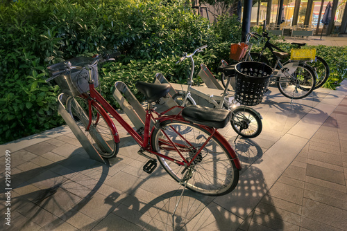 bicycles parked at metal outdoor rack during evening hours, modern paved stone walkway next to lush green hedge and building entryway, illuminated by bright overhead street or landscape lighting
