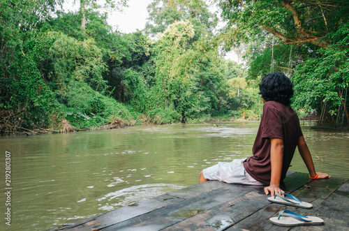 Fotografie A long-haired Southeast Asian male teenager with glasses sitting by the pier, re