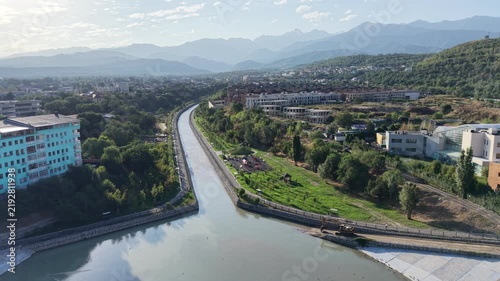 Aerial flyover above river park promenade toward mountains with luxury residential complexes at sunrise