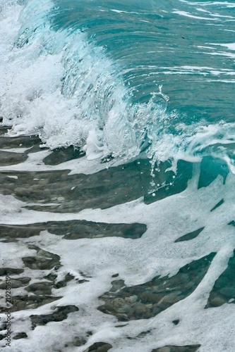 Fotografie A close-up action shot of a turquoise ocean wave crashing and breaking over smooth pebbles on a coastal beach, creating white sea foam and splashing water droplets