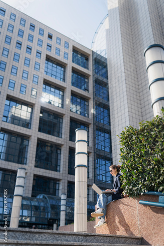 Millennial business woman working remotely on a laptop outdoors  sitting near a modern corporate building with a happy smile.
