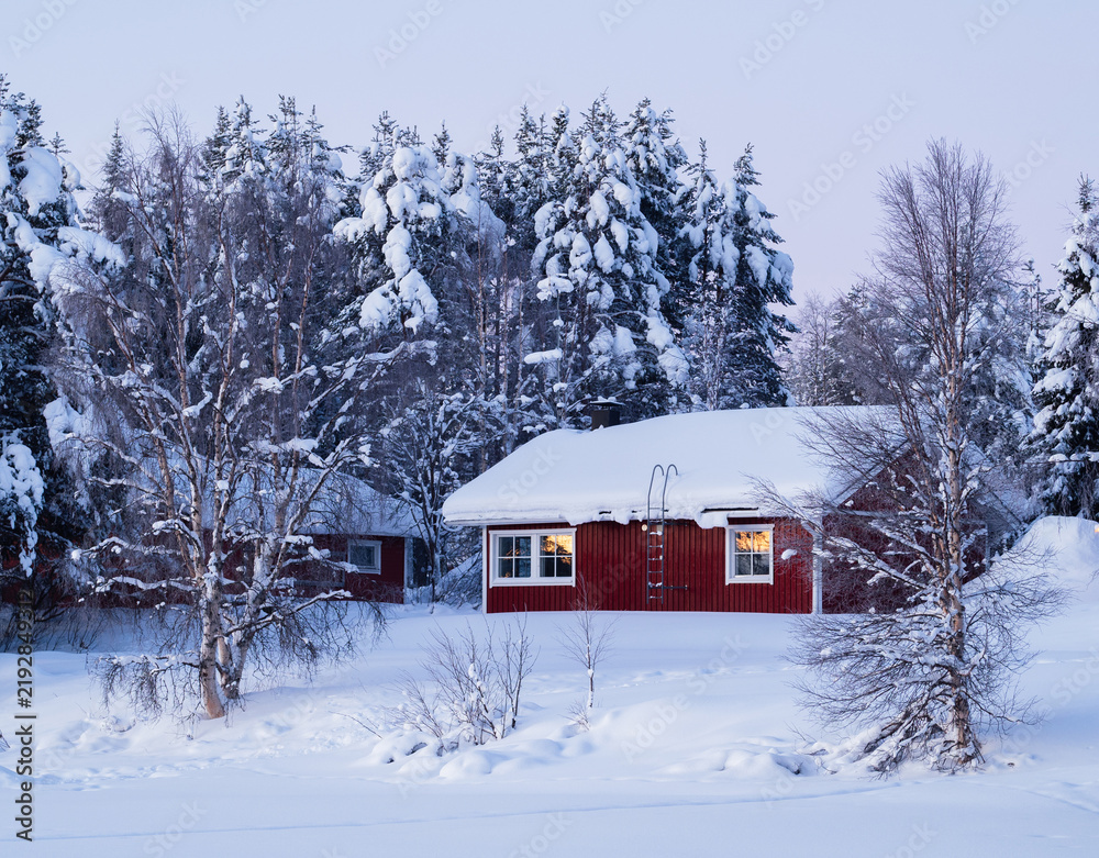 Obraz premium Traditional red finnish cabin in snowy winter forest with glowing window reflections
