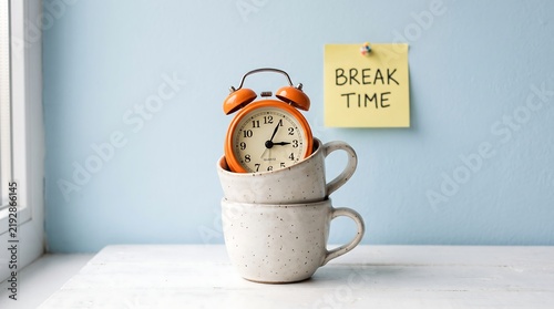 Orange alarm clock nestled in a ceramic mug with a break time sticky note on a windowsill