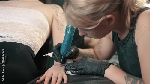 Professional tattoo artist creating a tattoo on a woman's forearm using a tattoo machine in a modern studio. Client relaxing during the tattooing procedure.
