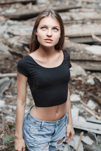 Candid portrait of a slender young woman with long brown hair  looking upwards with a thoughtful expression  wearing a black crop top and ripped denim shorts amidst decaying urban ruins and rubble.