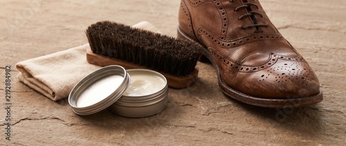 Brown leather brogue shoe and polishing brush alongside an open tin of conditioning wax and a soft cloth for traditional footwear maintenance
