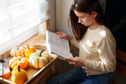 Teen girl reading book by window with pumpkins in autumn light