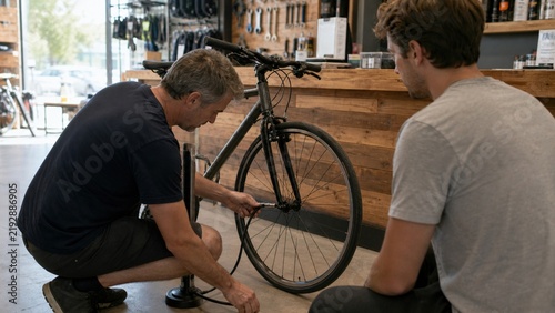 Mature bicycle mechanic and a younger customer inflating a front tire using a manual floor pump during a routine maintenance check inside a local retail repair shop