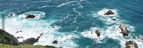 Фотография Turquoise ocean waves and foaming white surf surround rugged offshore rock formations below a wild coastal cliff, creating a dramatic natural seascape with rich marine textures