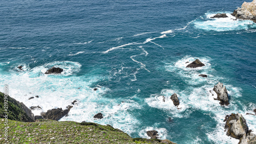 Принти на полотні Turquoise ocean waves and foaming white surf surround rugged offshore rock formations below a wild coastal cliff, creating a dramatic natural seascape with rich marine textures