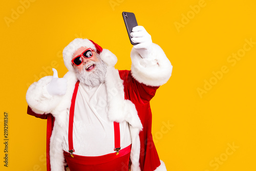 Fotografie Santa Claus taking a selfie with sunglasses and thumbs up in a red suit on a bri