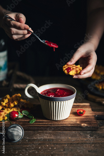 фотографія Hand dipping a mini waffle into a ceramic cup of homemade red berry jam on a dar