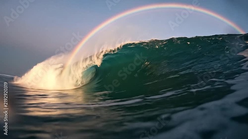 Large ocean wave breaking under bright rainbow arc.