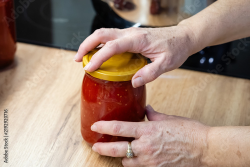 Hands closing yellow metal screw lid on glass jar of tomato sauce close up
