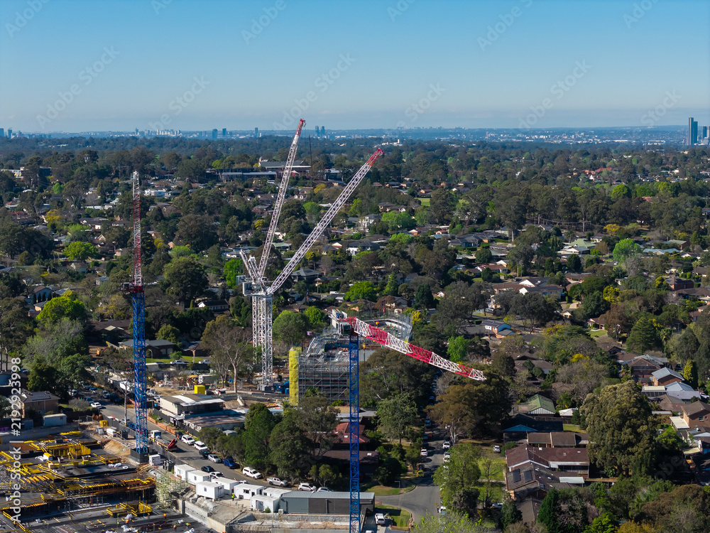 Obraz premium Construction cranes across a growing suburban Sydney district with established houses and distant skyline, illustrating redevelopment, housing growth and urban change
