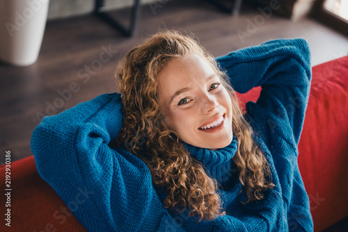 Konstfotografi Young woman with curly hair relaxing on a red sofa, wearing a blue knitted pullo