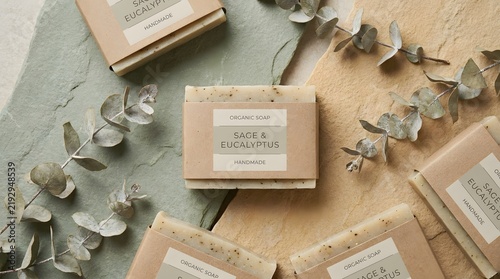 Several bars of handmade organic soap with sage and eucalyptus scents are arranged on green and beige textured stones. The soaps are labeled clearly and look fresh