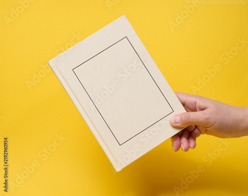 A person holding a closed light hardcover book with a frame on the cover against a yellow background