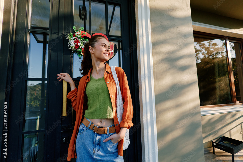Fototapeta premium Young woman standing outdoors near a building entrance, smiling and looking away. She wears a vibrant orange shirt and blue jeans. Lifestyle concept for urban fashion and leisure.