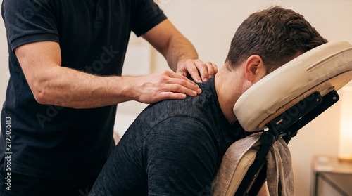 Masseur giving back massage to man sitting in massage chair. Therapist pressing shoulder muscles during treatment session in wellness studio.