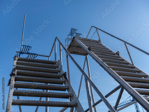 Galvanised steel stairs and a walkway platform rise against a blue sky, industrial access at a river shipyard.